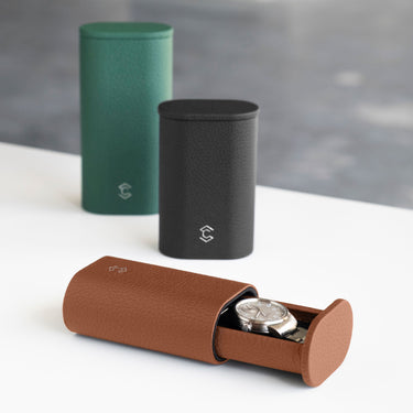 Lifestyle photo of open tan Oscar Watch roll holding a luxury watch with a metal bracelet in the foreground. Two closed, standing Oscar watch rolls are placed in the background in emerald and black leather. 